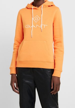 Person trägt einen leuchtend orangefarbenen GANT-Hoodie mit weißen Kordeln und schwarze Hose vor einem einfarbigen hellgrauen Hintergrund.