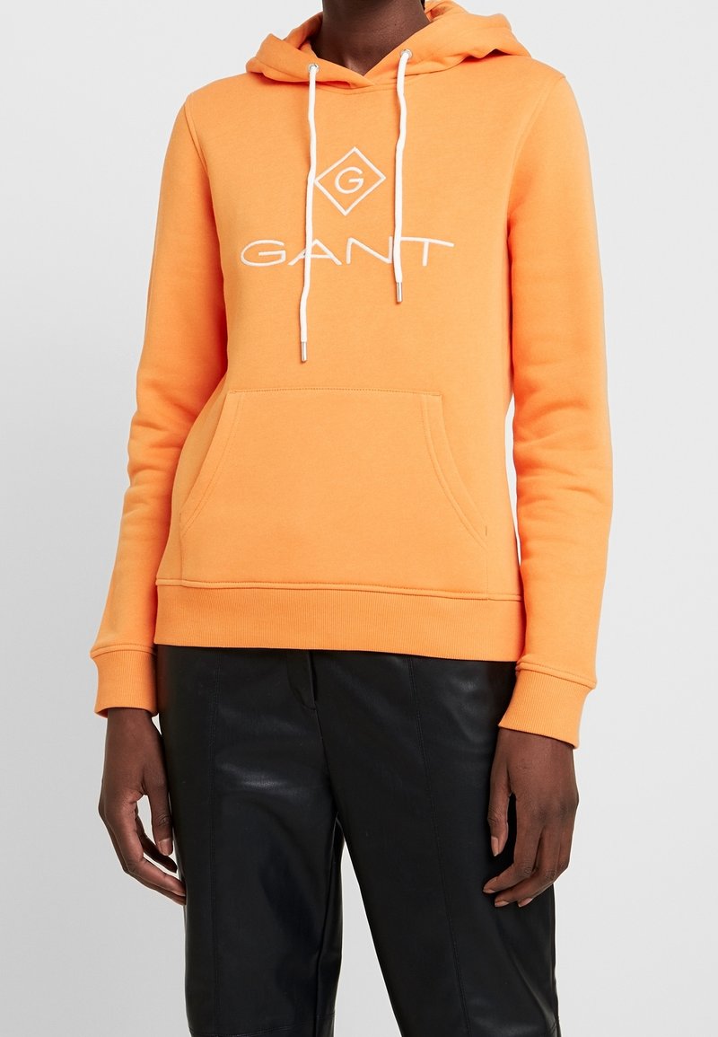 Personne portant un sweat à capuche GANT orange vif avec des cordons blancs et un pantalon noir sur un fond gris clair uni.