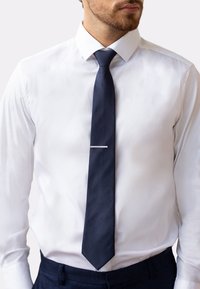 Cravate en soie bleu marine avec une texture lisse, ornée d'un clip de cravate en argent. Portée avec une chemise habillée blanche et un pantalon sombre. Design slim et allongé.
