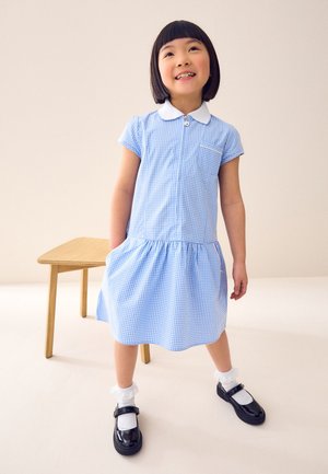 Niña joven con vestido de cuadros azul con cuello blanco, calcetines blancos y zapatos negros, de pie junto a una mesa de madera contra un fondo liso.