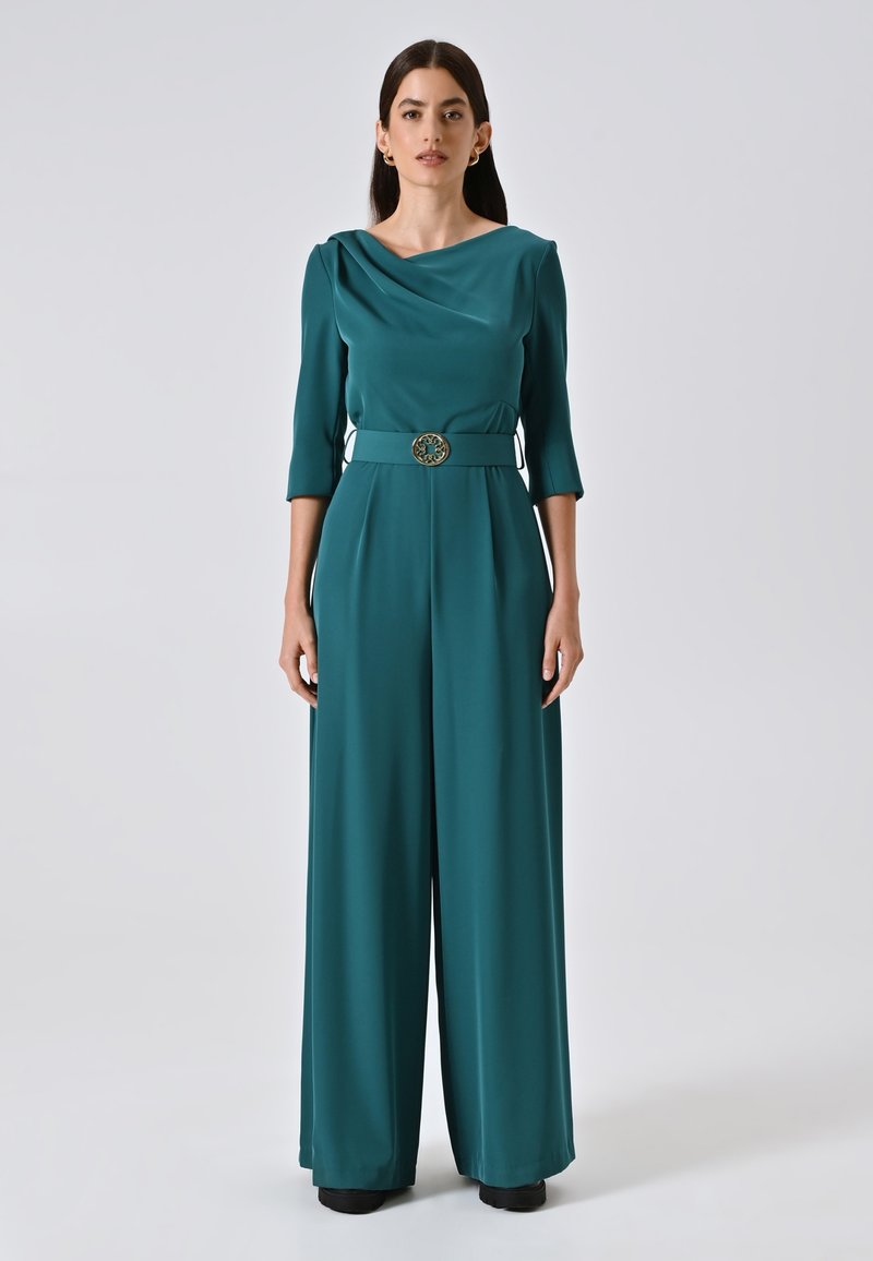 Tuta teal con scollatura drappeggiata, maniche a tre quarti, vita cintrata con fibbia decorativa, design a gamba larga, tessuto liscio.