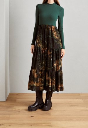 Femme portant un haut vert à manches longues avec une jupe longue patchwork fluide marron et verte et des bottes plateforme épaisses foncées, debout sur un sol en bois.