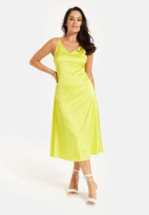 Sleeveless yellow midi dress with a V-neckline, smooth texture, and flared silhouette. Worn with white lace-up sandals.