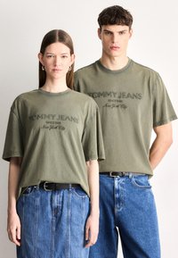Tricouri din bumbac verde măsliniu cu logo-ul brodat "Tommy Jeans", asortate cu blugi albaștri din denim. Croială lejeră și mâneci scurte pentru ambele modele.