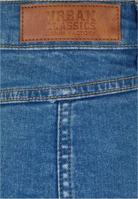 Modrá džínová tkanina s viditelným šitím, na pásu s hnědým koženým štítkem s nápisem "URBAN CLASSICS DENIM FACTORY".