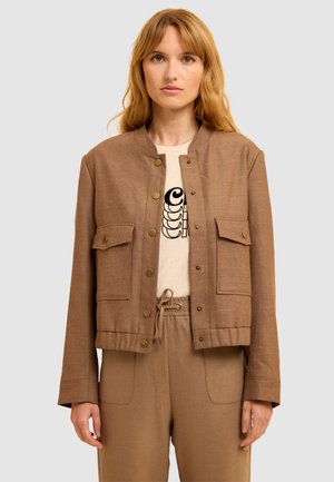 Frau, die eine braune, geknöpfte Jacke über einem beigen Shirt mit Grafik und passenden braunen Jogginghosen trägt, vor einem schlichten, hellen Hintergrund.