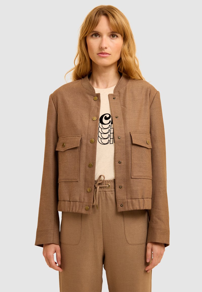 Femme portant une veste marron à boutons sur un t-shirt graphique beige et un pantalon à cordon de serrage marron assorti, sur un fond clair et uni.