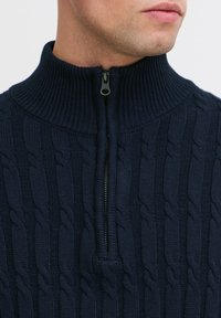 Suéter de punto azul marino con un alto cuello acanalado. Cuenta con un cierre de media cremallera en el frente, patrón de trenzado y textura suave.