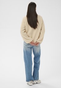 Femme debout, dos tourné vers la caméra, portant une veste polaire beige, un jean bleu et des baskets blanches, les mains jointes dans le dos.