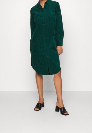 Personne portant une robe chemise en velours côtelé vert foncé à manches longues avec des chaussures noires à talons blocs ouverts, debout sur un sol blanc.