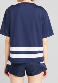 Granatowa koszulka sportowa z krótkim rękawem, z białymi siateczkowymi paskami u dołu. W połączeniu z błyszczącymi granatowymi szortami, które prezentują sportowy wygląd.