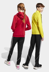 Červená mikina s bielymi pruhmi, čierne nohavice a žltá košeľa s dlhým rukávom. Obe osoby majú na sebe tenisky s kontrastnými akcentmi. Pohľad zozadu.