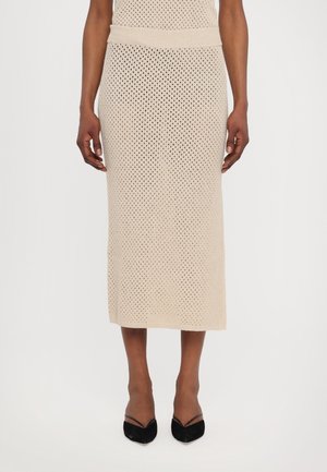 Beige strikket midi-nederdel med perforeret mønster båret af person med mørk hud, kombineret med sorte spidse hæle med tynde stropper.