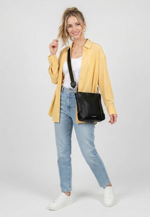 Ung kvinde iført gul skjorte, hvid top, lyseblå jeans, hvide sneakers og sort crossbody-taske, smilende og poserende foran en grå baggrund.