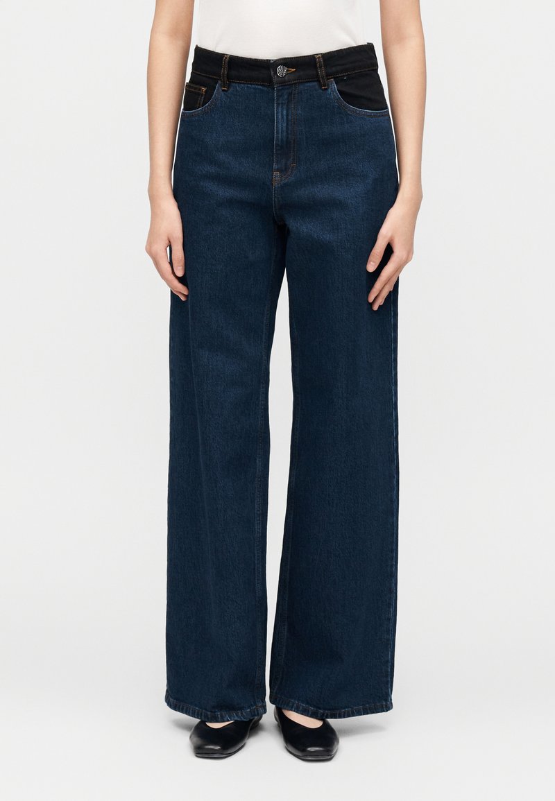 Τζιν παντελόνια με φαρδιά μπατζάκια σε σκούρο μπλε χρώμα και ψηλή μέση, με μαύρες τσέπες, ορατή ραφή και κλείσιμο με κουμπιά μπροστά.