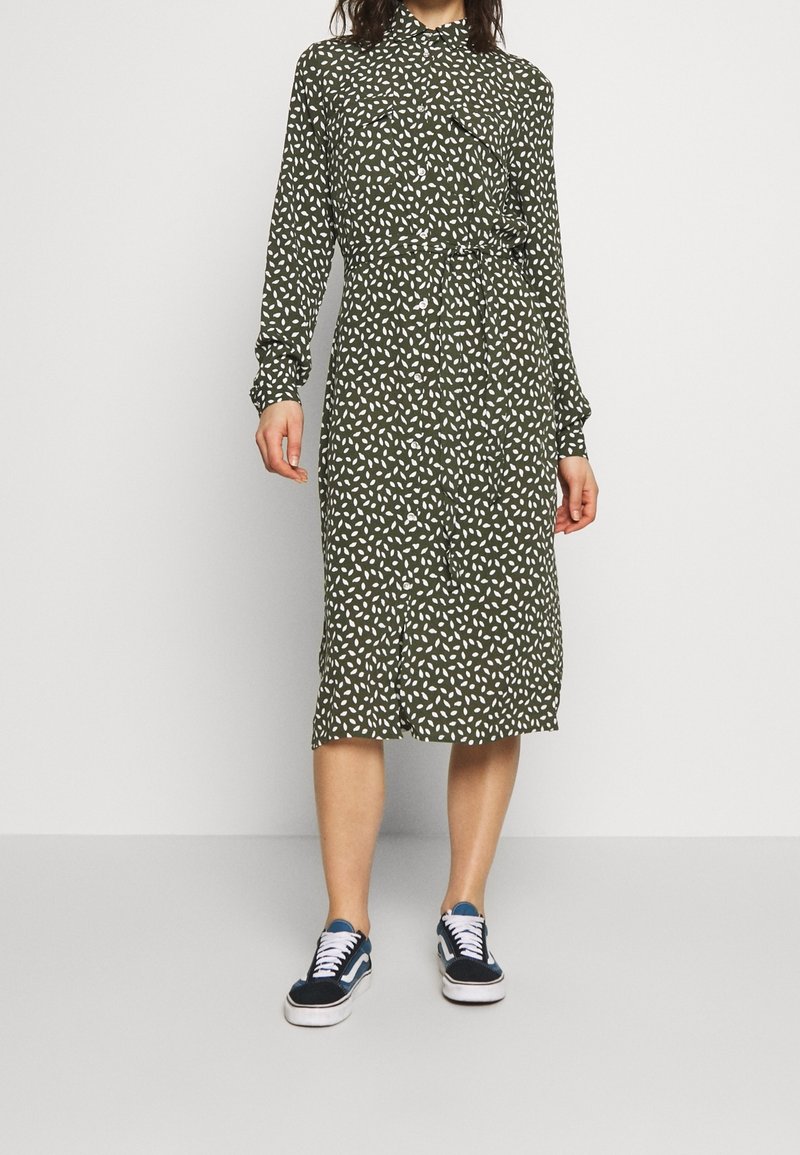 Femme portant une robe midi verte à pois blancs et manches longues, associée à des baskets noires et blanches.