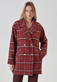 Donna che indossa un cappotto doppiopetto a quadri rosso e grigio con bottoni argentati, in posa con una mano in tasca su uno sfondo neutro.
