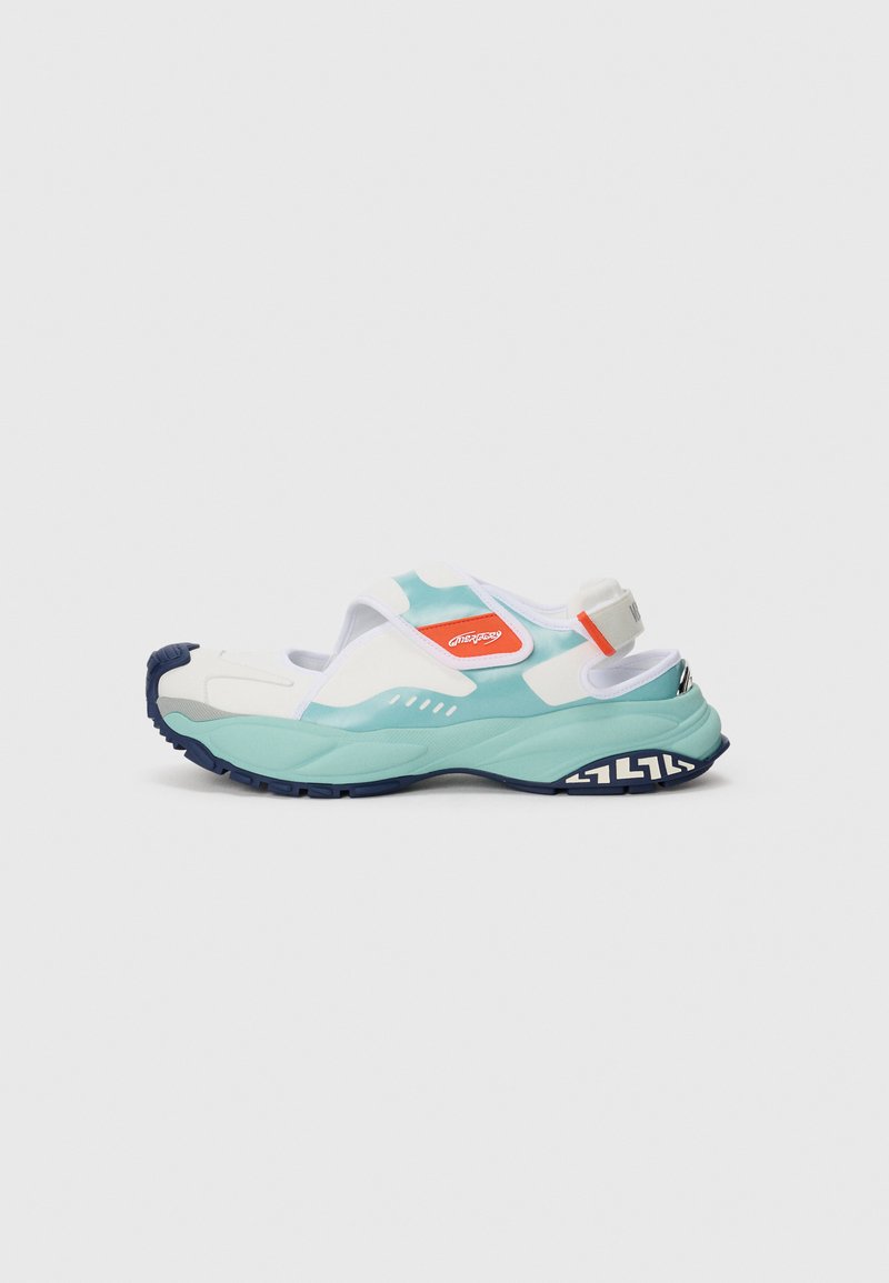 White, teal, and navy slip-on sneaker with an open heel, orange logo patch, and a thick patterned sole on a light background.