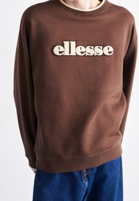 Hnědá mikina s vyšitým logem "ellesse" v krémové barvě, s ribovaným výstřihem a manžetami, v kombinaci s modrými džíny.