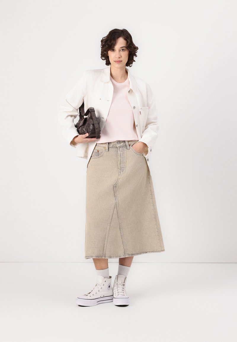 Femme en veste crème, chemise rose, jupe midi beige, baskets montantes blanches, tenant un sac à main sombre devant un fond blanc uni.