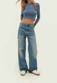 Kvinna som bär en blå långärmad croptop, högmidjade vida slitna jeans och vita sneakers, står med händerna bakom ryggen.