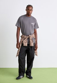 Jongeman staat op groene vloer, draagt een grijs bedrukt T-shirt, zwarte broek, een geruite shirt om zijn middel geknoopt, en zwarte sneakers.