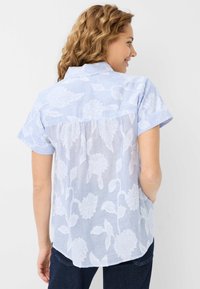 Korte mouw, lichtblauw shirt met witte bloemenpatronen, zachte textuur en een plooi detail aan de achterkant. Gebogen zoom met een ontspannen pasvorm.