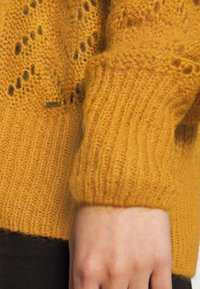 Mosterdgele gebreide trui met een textuurpatroon, geribbelde boorden en een klein merklabel nabij de zoom. Zacht en lichtgewicht materiaal.