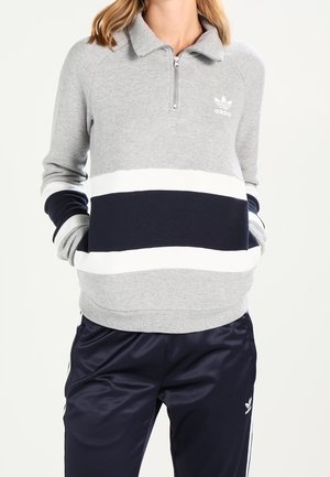 Osoba ubrana w szarą i granatową bluzę Adidas z białymi paskami oraz granatowe dresy Adidas na jednolitym białym tle.