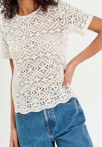 Spitzenoberteil in Off-White mit floralen Mustern, kurzen Ärmeln, welligem Saum und lockerer Passform, kombiniert mit blauen Jeans.