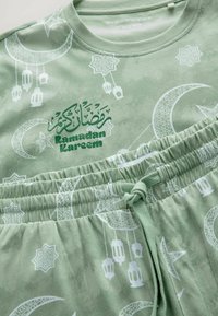 Green nightwear set featuring a crescent moon and lantern pattern. Text "Ramadan Kareem" printed in darker green on the top. Soft material.