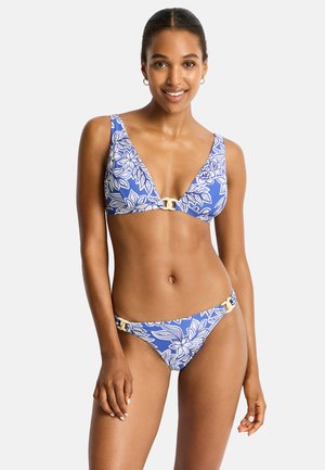 Donna sorridente che indossa un bikini floreale blu e bianco con dettagli ad anello sopra e sotto su uno sfondo chiaro uniforme.