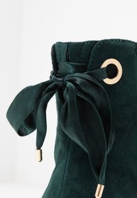 Stivaletto alla caviglia in suede verde scuro con una finitura testurizzata, caratterizzato da un nastro di satin legato a fiocco e un dettaglio in metallo dorato.