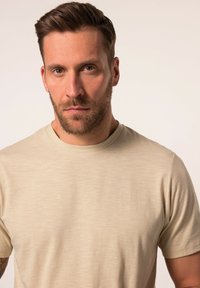 Camiseta de manga corta beige, hecha de tela texturizada, con un sutil logo en relieve en el pecho y un cuello redondo.