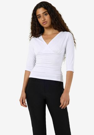 Femme aux longs cheveux bouclés portant un haut blanc à col en V avec des manches trois-quarts et un pantalon noir sur un fond uni.
