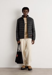 Zwarte gewatteerde pufferjas met een hoge kraag, een beige sweatshirt eronder, crèmekleurige broek en een zwarte totebag met rode accenten. Bruine schoenen.