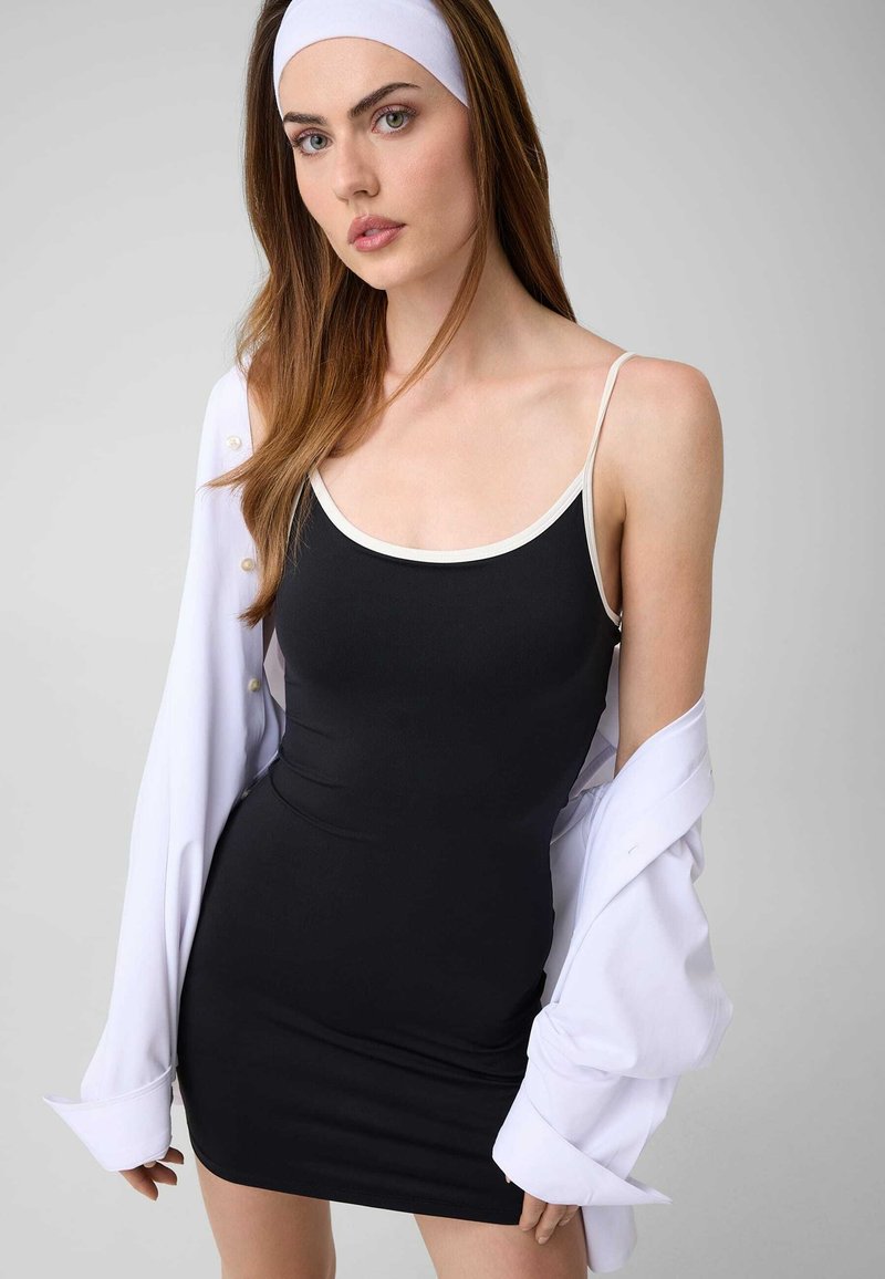 Femme aux longs cheveux bruns porte une robe noire sans manches avec un bord blanc, un bandeau blanc et une chemise blanche boutonnée partiellement drapée.