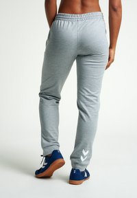 Grijze sweatpants met een elastische tailleband, twee zijzakken en een wit chevronlogo op het onderste deel van het linkerpijpen. Gecombineerd met blauwe sneakers.