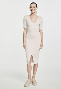Sukienka midi dziewiarska w jasnym beżu z krótkimi rękawami, dekoltem w kształcie litery V, guzikami z przodu i rozcięciem po boku. Połączona z jasnymi butami na platformie.