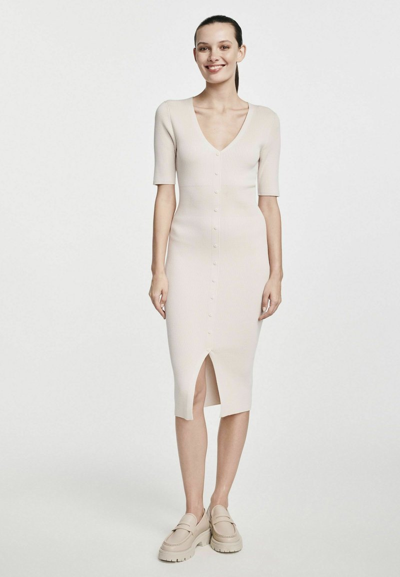 Sukienka midi dziewiarska w jasnym beżu z krótkimi rękawami, dekoltem w kształcie litery V, guzikami z przodu i rozcięciem po boku. Połączona z jasnymi butami na platformie.
