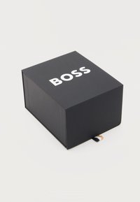 Μαύρο ορθογώνιο κουτί με τη λέξη "BOSS" σε λευκό στο επάνω μέρος και μια μικρή υφασμάτινη ετικέτα σε μαύρο, λευκό και μπεζ στη βάση.