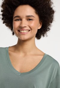 T-shirt verde chiaro con scollo a V e una consistenza morbida. Il modello mostra il taglio rilassato della maglietta e il collo, con maniche corte e tessuto morbido.