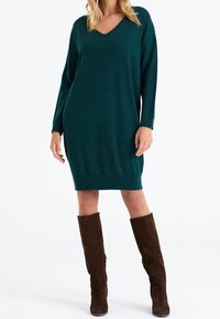 Robe en maille bleu sarcelle avec un col en V, des manches longues et une coupe décontractée, se terminant juste au-dessus du genou. Associée à des bottes hauteur genou en suède marron.