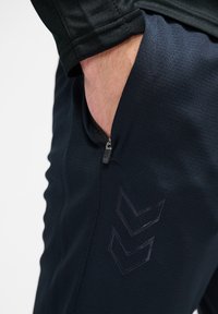 Marineblauwe sportbroek met een ritszak, gestoffeerd materiaal en een subtiele chevronlogo in een donkere tint aan de zijkant.