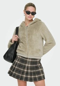 Beige Kunstfell-Jacke mit Reißverschluss und Kapuze, kombiniert mit einem karierten, plissierten Rock in Schwarz, Creme und Grau, sowie einer schwarzen Lederschultertasche.