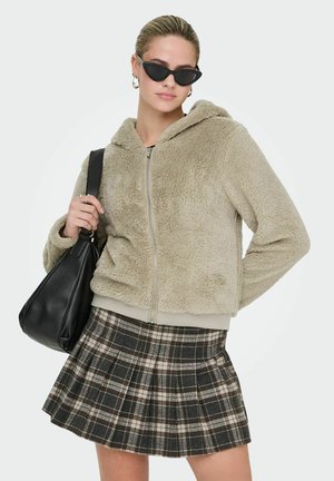Beige Kunstfell-Jacke mit Reißverschluss und Kapuze, kombiniert mit einem karierten, plissierten Rock in Schwarz, Creme und Grau, sowie einer schwarzen Lederschultertasche.