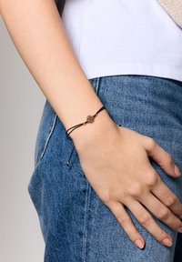 Czarna regulowana bransoletka z cordu z złotym kwiatowym charms, z dwoma złotymi koralikami jako akcentami, noszona na ręce na tle dżinsowego materiału.