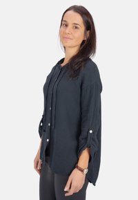 Navyblaue Bluse mit Knopfleiste aus strukturiertem Stoff, mit Kapuze, Kordelzügen und hochgerollten Ärmeln mit Knopfakzenten. Tragen Sie dazu schwarze Leggings.