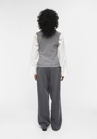 Un gilet gris à côtes sur une chemise blanche à manches longues, associé à un pantalon gris à jambes larges. Texture lisse, design superposé et couleurs douces.