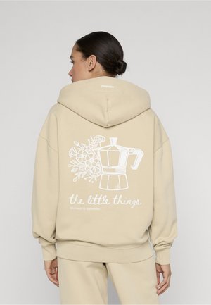 Femme aux cheveux bruns attachés en chignon portant un sweat à capuche et un pantalon beiges, le dos du sweat montre une illustration d'une cafetière blanche et une fleur avec le texte « les petites choses ».
